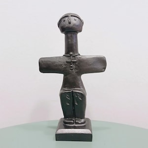 May include: A dark bronze-colored sculpture of a stylized human figure. The figure has a head, a long neck, outstretched arms, and a cross-shaped pendant. The sculpture is mounted on a square base and stands on a light green surface.