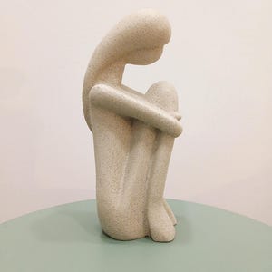 May include: A beige sculpture of a person in a seated position, with knees drawn up and head resting on them. The figure has a textured surface and is placed on a light green table. The background is a plain white wall.