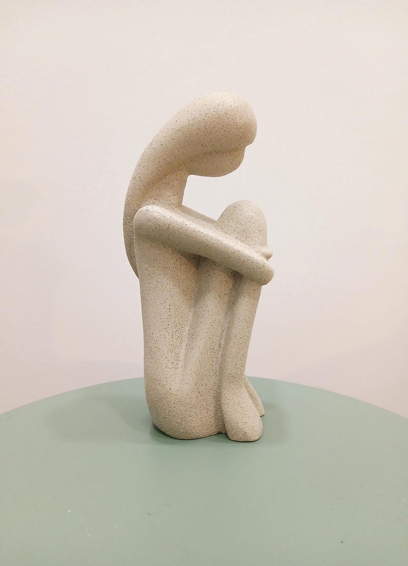May include: A beige sculpture of a person in a seated position, with knees drawn up and head resting on them. The figure has a textured surface and is placed on a light green table. The background is a plain white wall.