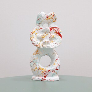 May include: A white, abstract sculpture with a stacked design. The sculpture features three circular forms, each with a hole in the center. It is decorated with splatters of red, yellow, and blue paint. The sculpture is on a light green surface.