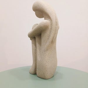 May include: A light beige sculpture of a person in a seated position, with their knees drawn up and arms wrapped around them. The sculpture has a textured surface and sits on a light green surface.