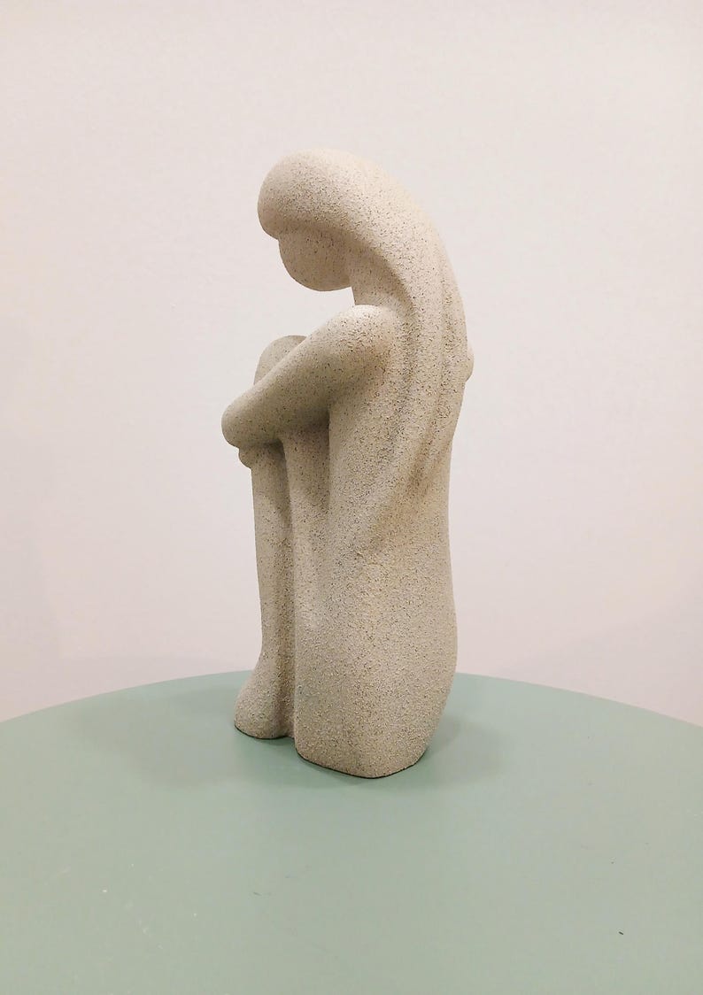 May include: A light beige sculpture of a person in a seated position, with their knees drawn up and arms wrapped around them. The sculpture has a textured surface and sits on a light green surface.