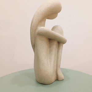 May include: A beige-colored sculpture of a seated figure, knees drawn up to the chest, arms wrapped around the legs. The figure is on a light green surface, with a neutral background. The sculpture has a textured surface.