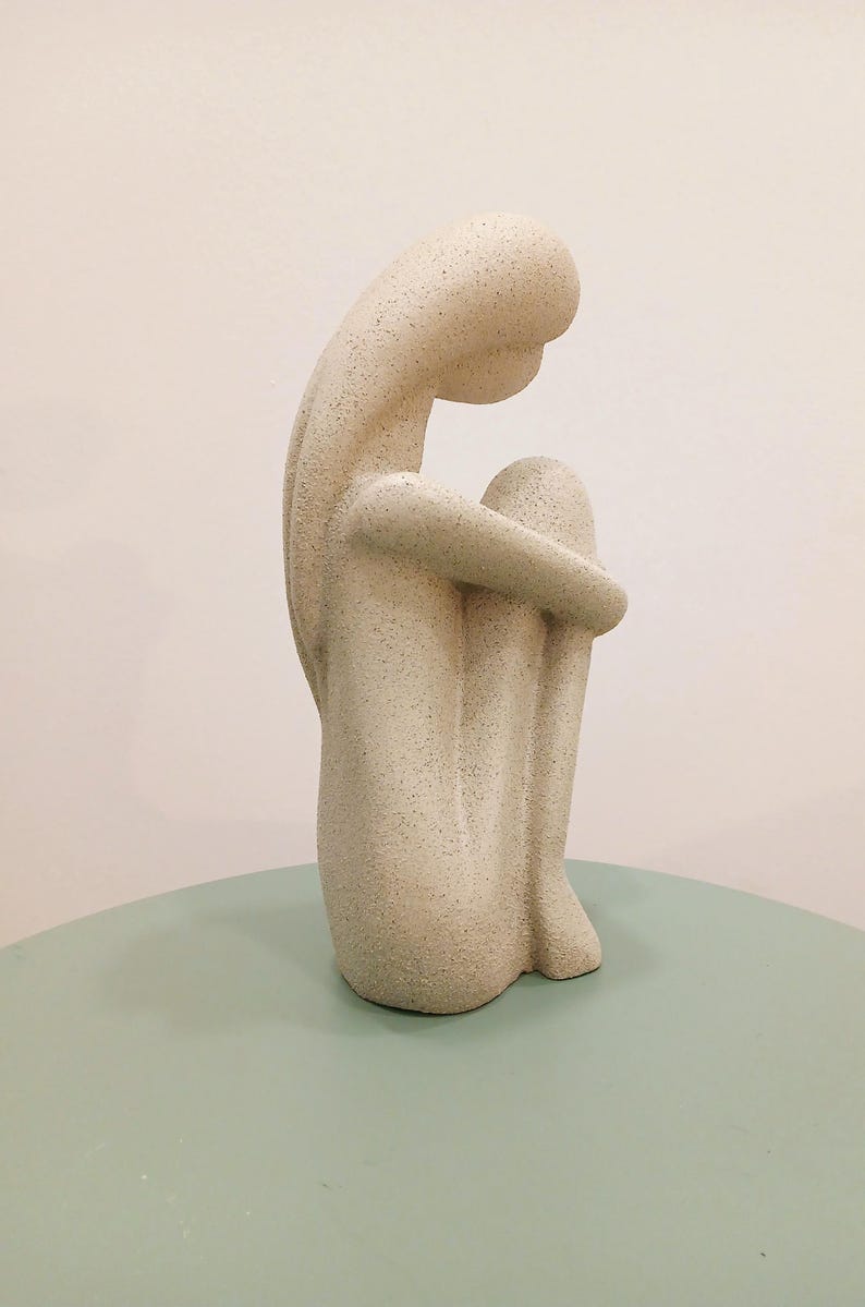 May include: A beige-colored sculpture of a seated figure, knees drawn up to the chest, arms wrapped around the legs. The figure is on a light green surface, with a neutral background. The sculpture has a textured surface.