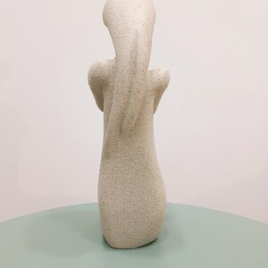 May include: A light beige abstract sculpture with a textured surface. The sculpture has a slender, elongated form with a rounded head and arms that curve inward. The sculpture is displayed on a light green surface.