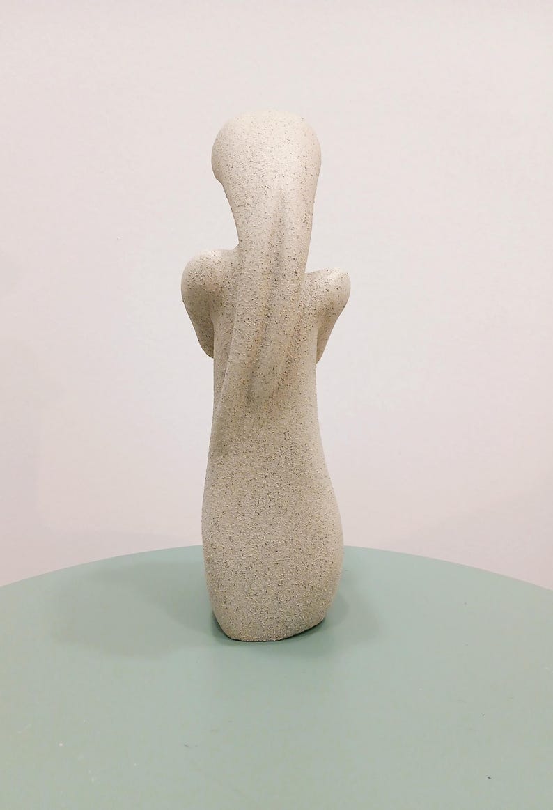 May include: A light beige abstract sculpture with a textured surface. The sculpture has a slender, elongated form with a rounded head and arms that curve inward. The sculpture is displayed on a light green surface.
