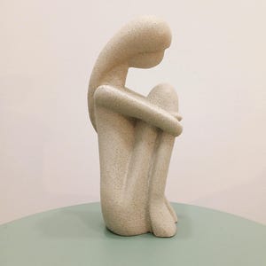 May include: A beige, abstract sculpture of a seated figure with arms wrapped around the knees. The sculpture has a textured surface and is positioned on a light green table. The figure's head is bowed, suggesting contemplation or introspection.