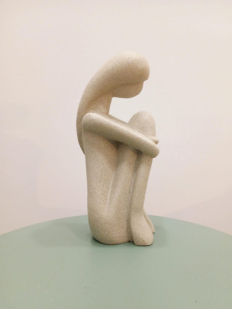 May include: A beige, abstract sculpture of a seated figure with arms wrapped around the knees. The sculpture has a textured surface and is positioned on a light green table. The figure's head is bowed, suggesting contemplation or introspection.