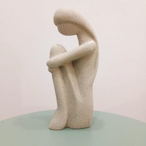 May include: A beige, textured sculpture of a person in a seated position, with arms wrapped around the knees. The figure has a smooth, rounded head and elongated body. The sculpture is on a light green surface.