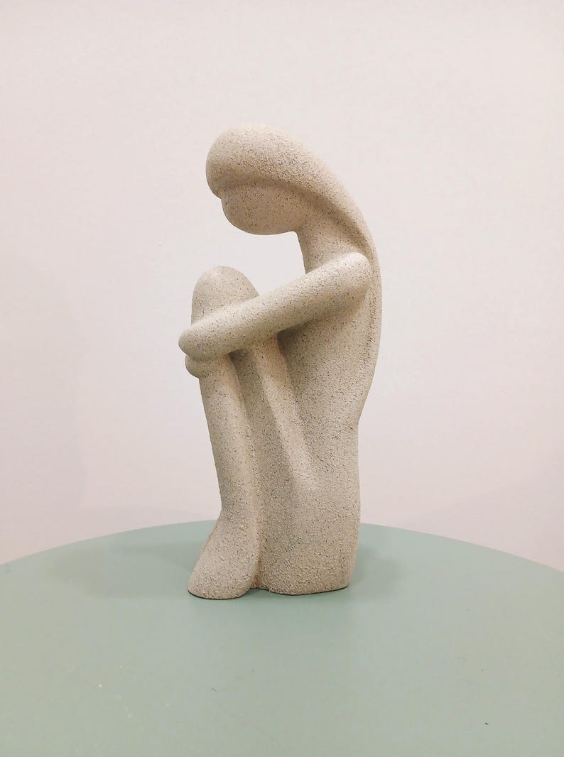May include: A beige, textured sculpture of a person in a seated position, with arms wrapped around the knees. The figure has a smooth, rounded head and elongated body. The sculpture is on a light green surface.