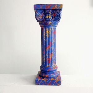May include: A decorative column featuring a classic Ionic capital with volutes and a fluted shaft. The entire structure is painted in a vibrant blue base colour, accented with a chaotic, multi-coloured paint splatter pattern in shades of red and yellow.