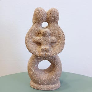 Pode incluir: Uma escultura de cerâmica bege de uma família. A escultura representa duas figuras de pé de costas uma para a outra, com uma figura menor de pé no espaço entre elas. A escultura tem uma superfície áspera e texturizada.