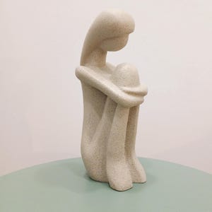 May include: A beige-colored sculpture of a seated figure, with the head resting on the knees. The figure is in a curled position, with arms wrapped around the legs. The sculpture is on a light green surface.