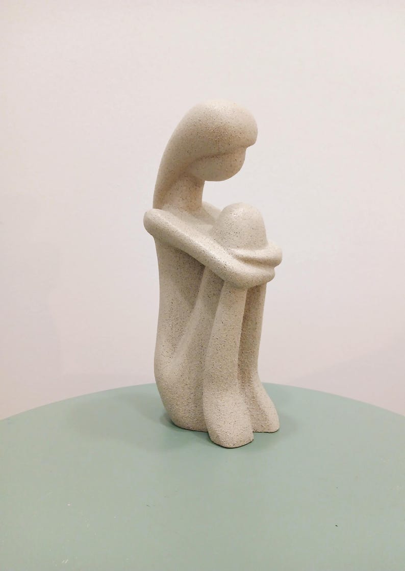 May include: A beige-colored sculpture of a seated figure, with the head resting on the knees. The figure is in a curled position, with arms wrapped around the legs. The sculpture is on a light green surface.