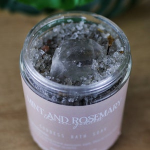 Puede incluir: Un tarro de vidrio transparente lleno de sales de baño y un cristal transparente. El tarro tiene una etiqueta rosa que dice "Mint and Rosemary Goddess Bath Soak".