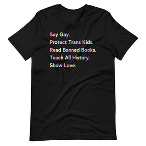 May include: Black t-shirt with a message in rainbow colours that reads: "Say Gay. Protect Trans Kids. Read Banned Books. Teach All History. Show Love."