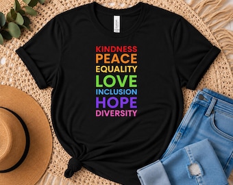 May include: A black short-sleeved t-shirt featuring a vertical list of words printed in rainbow colors: KINDNESS in red, PEACE in orange, EQUALITY in yellow, LOVE in green, INCLUSION in blue, HOPE in purple, and DIVERSITY in pink.