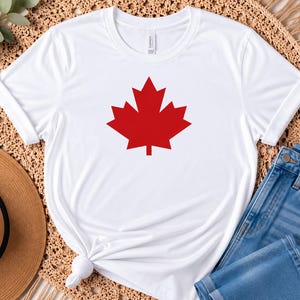 Puede incluir: Una camiseta blanca de manga corta con un gr&aacute;fico grande de una hoja de arce roja s&oacute;lida centrado en el pecho. La prenda tiene un cuello redondo y est&aacute; confeccionada con un tejido liso, mostrando un dise&ntilde;o limpio con un s&iacute;mbolo ic&oacute;nico.