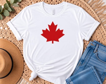 Pode incluir: Uma t-shirt branca de manga curta com um gráfico grande de uma folha de ácer vermelha sólida centrado no peito. A peça tem um decote redondo e é feita de tecido liso, exibindo um design limpo com um símbolo icónico e marcante.