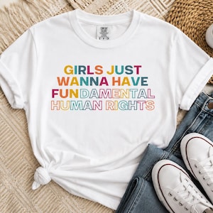 May include: A white short-sleeved t-shirt featuring the multicolored text GIRLS JUST WANNA HAVE FUN DAMENTAL HUMAN RIGHTS printed on the front. The shirt has a crew neckline and a small brand label visible at the inner collar area.