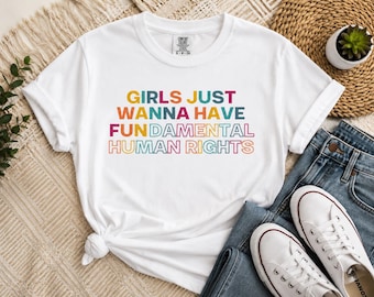 Puede incluir: Una camiseta blanca de manga corta con el texto multicolor GIRLS JUST WANNA HAVE FUN DAMENTAL HUMAN RIGHTS impreso en la parte delantera. La camiseta tiene un cuello redondo y una pequeña etiqueta de marca visible en el cuello.