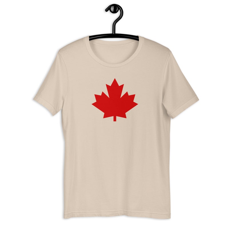 Puede incluir: Una camiseta de manga corta de color beige con un gr&aacute;fico de una hoja de arce roja s&oacute;lida centrado en el pecho. La prenda se muestra en una percha de pl&aacute;stico negra, con cuello redondo, corte est&aacute;ndar, mangas cortas y dobladillo recto.