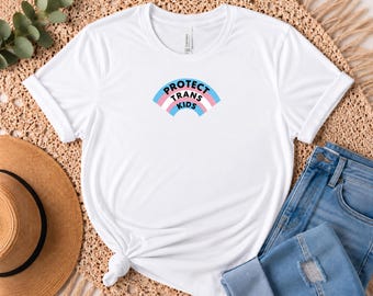 May include: A white short-sleeved t-shirt featuring a graphic of an arched rainbow with horizontal stripes in light blue, pink, and white. The text "PROTECT TRANS KIDS" is printed in black capital letters across the center of the rainbow.