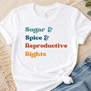 May include: A white short-sleeve t-shirt featuring the text Sugar & Spice & Reproductive Rights printed in four distinct colors: teal, blue, red, and orange. The shirt has a crew neckline and is displayed with blue denim jeans.