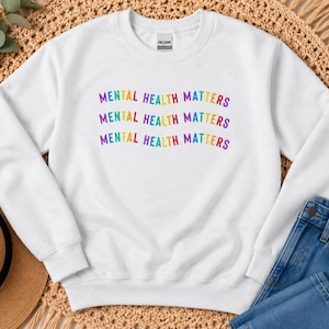 May include: A white crewneck sweatshirt featuring the text MENTAL HEALTH MATTERS printed three times in a wavy, multi-colored font. The design uses purple, blue, green, yellow, orange, and red letters. The garment has long sleeves and a ribbed collar.