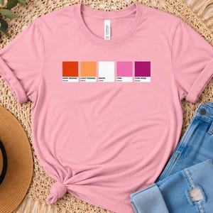 May include: A light pink short-sleeve t-shirt featuring a graphic of five rectangular color swatches in dark orange, light orange, white, pink, and dark rose, each labeled with its respective color name in small black text underneath.