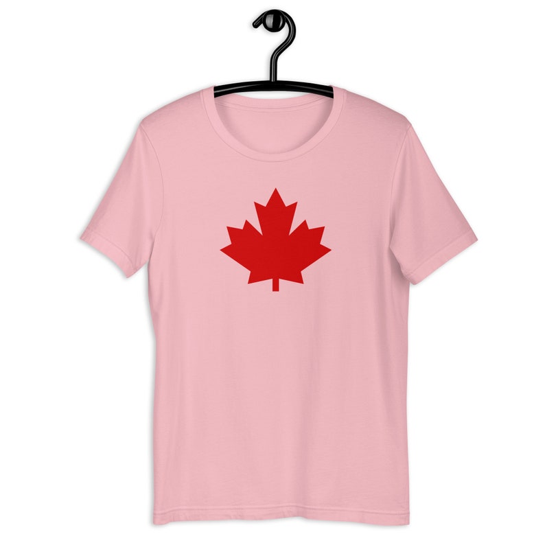 Puede incluir: Una camiseta de manga corta de color rosa claro con un gr&aacute;fico de hoja de arce roja s&oacute;lida centrado en el pecho. La prenda se muestra en una percha de pl&aacute;stico negra, con un cuello redondo y un dise&ntilde;o de dobladillo recto est&aacute;ndar.
