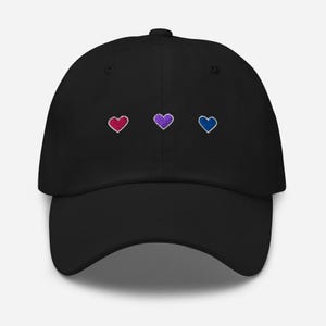 Peut inclure: Casquette de baseball noire avec trois cœurs brodés en rouge, violet et bleu. Les cœurs sont alignés horizontalement sur le devant. La casquette a une visière incurvée et un bouton sur le dessus.