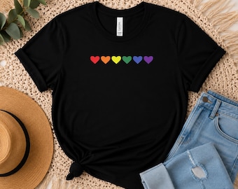 May include: A black short-sleeved t-shirt featuring a horizontal row of six small hearts in rainbow colors across the chest. The shirt has a crew neckline and a standard fit, with a white brand label visible on the inside of the collar.