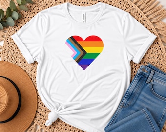 May include: A white short-sleeve t-shirt featuring a heart-shaped graphic on the chest. The heart is filled with the Progress Pride flag design, incorporating horizontal stripes of red, orange, yellow, green, blue, purple, black, brown, light blue, pink, and white.