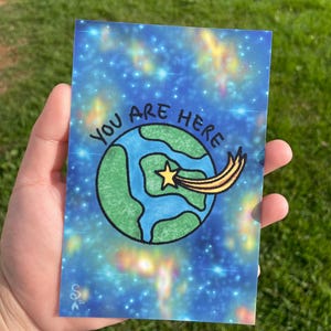 May include: A rectangular greeting card with a blue galaxy background and the words "YOU ARE HERE". The card features a green and blue Earth with a shooting star. The card is held in a person's hand against a background of green grass.