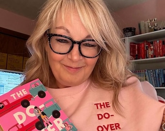 Puede incluir: Una mujer con una sudadera rosa con las palabras "THE DO-OVER" bordadas en rojo. Sostiene un libro con el mismo título, con ilustraciones de camiones y personas. Lleva gafas con montura negra.