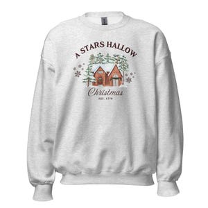May include: Light grey sweatshirt with a graphic of a brick house covered in snow, with the text "A Stars Hallow Christmas" and "EST. 1779". The design includes snowflakes and evergreen trees, creating a festive holiday theme.