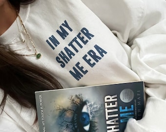 Pode incluir: Uma camisa de moletom branca com o texto "In My Shatter Me Era" impresso em azul. Uma pessoa está segurando um livro intitulado "Shatter Me" com um gráfico de olho azul na capa.