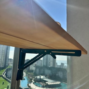 May include: A light brown wooden folding table with dark green metal supports. The table is mounted on a balcony railing, with a city skyline and blue sky visible in the background.