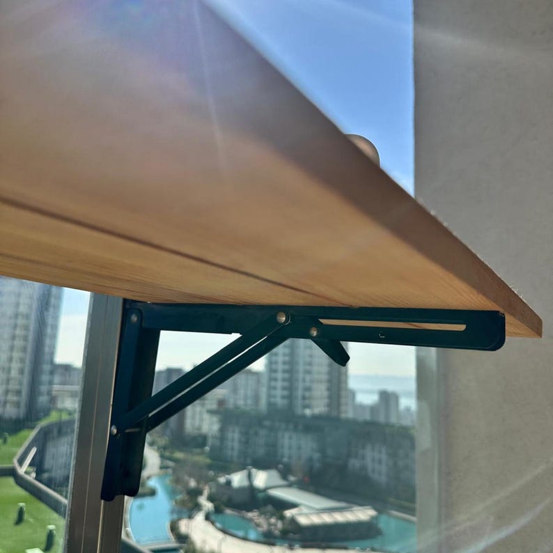 May include: A light brown wooden folding table with dark green metal supports. The table is mounted on a balcony railing, with a city skyline and blue sky visible in the background.