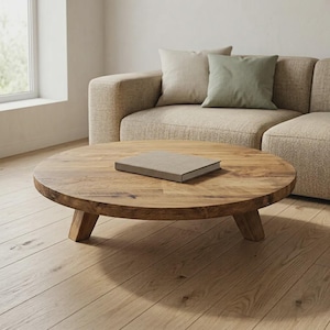 Pode incluir: Uma mesa de centro redonda e baixa feita de madeira natural com um padrão de grão visível, apoiada por três pernas curtas e robustas. Um único livro retangular com uma capa lisa de cor clara repousa plano no centro do tampo da mesa.