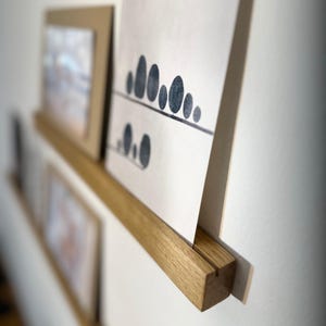 Może przedstawiać: Drewniana półka na zdjęcia zamontowana na białej ścianie, prezentująca oprawione grafiki i wydruki. Półka wykonana jest z jasnego drewna i mieści kilka dzieł sztuki, w tym wydruk z czarno-białym wzorem drzewa.