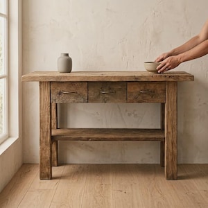 Può includere: Una consolle rustica in legno con tre cassetti e un ripiano inferiore. Il tavolo mostra venature e nodi naturali del legno. Un piccolo vaso grigio e una ciotola sono sul tavolo. Il tavolo misura circa 152 cm di lunghezza, 41 cm di larghezza e 76 cm di altezza.