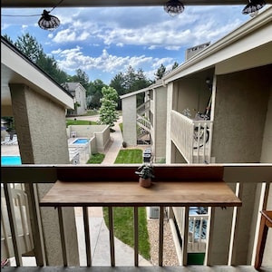 May include: View from a balcony with a wooden table attached to the railing. A small potted plant sits on the table. The background shows a courtyard with a pool, buildings, and a cloudy sky. String lights hang overhead.