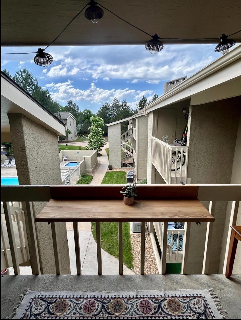 May include: View from a balcony with a wooden table attached to the railing. A small potted plant sits on the table. The background shows a courtyard with a pool, buildings, and a cloudy sky. String lights hang overhead.
