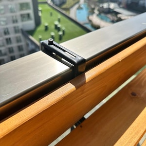 May include: A close-up of a black plastic bracket securing a stainless steel railing to a wooden deck. The wooden deck has a natural wood grain finish. The background shows a view of a building and a green area.