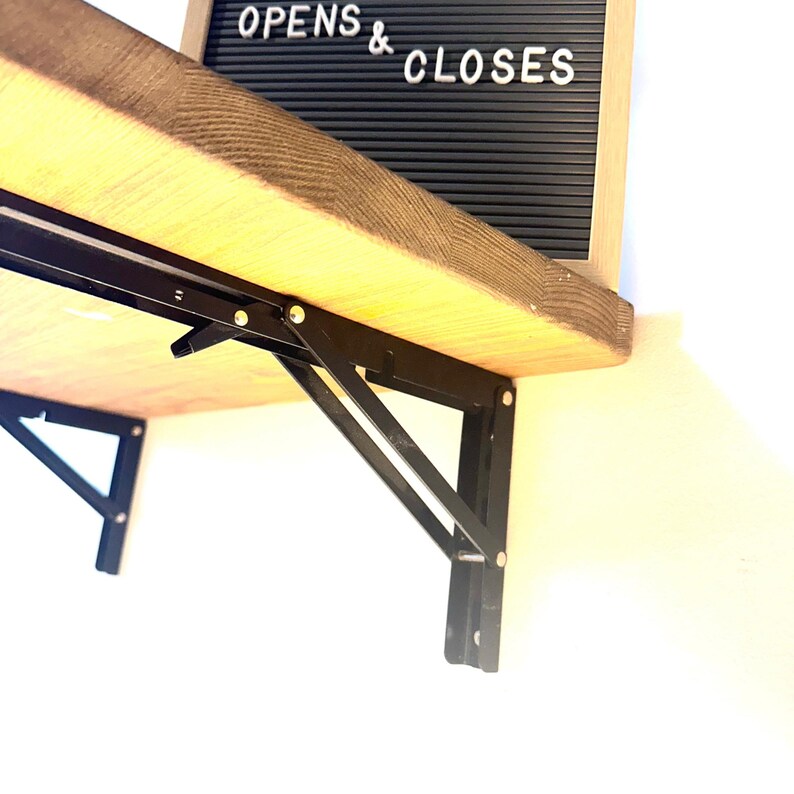 May include: A light brown wooden shelf mounted with black metal folding brackets. A black letter board with white text reading OPENS & CLOSES is positioned on the shelf. The brackets feature a triangular support design with visible rivets.