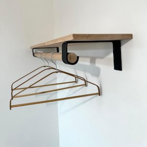 May include: A wooden shelf with a black metal rod and brackets, designed for hanging clothes. Three gold-coloured wire hangers are suspended from the rod. The shelf is mounted on a white wall.