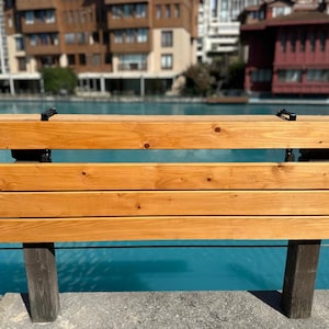 May include: A wooden bench with a light brown finish. The bench has three horizontal planks for seating and a backrest. The supports are dark brown. The background features a body of water and buildings.