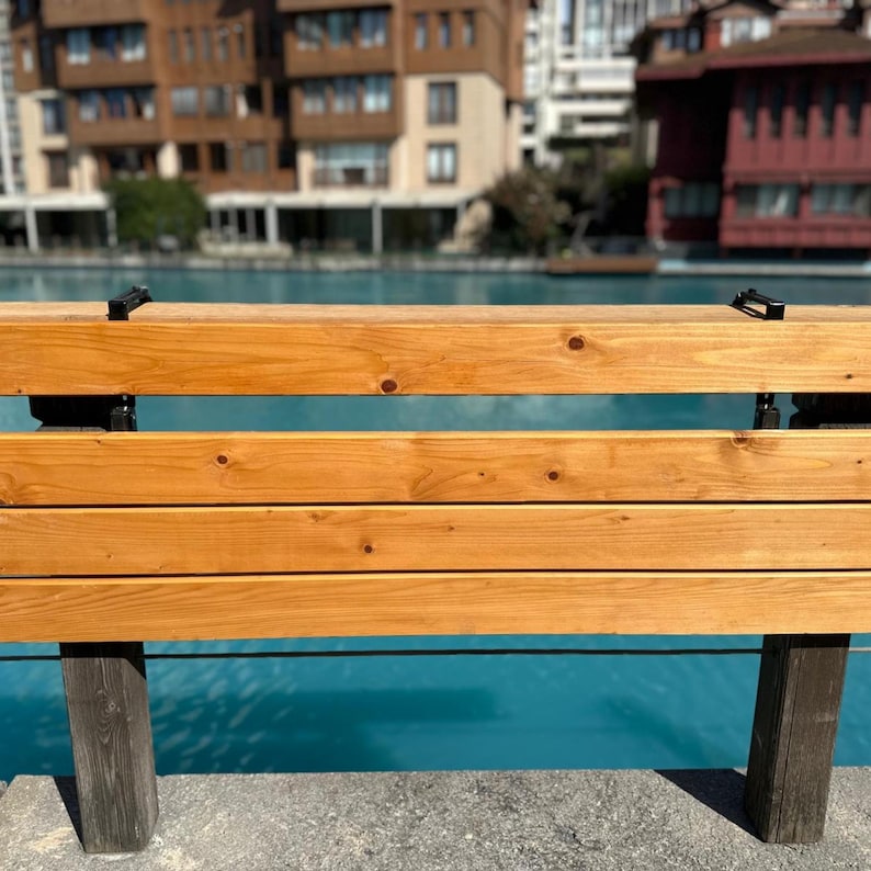 May include: A wooden bench with a light brown finish. The bench has three horizontal planks for seating and a backrest. The supports are dark brown. The background features a body of water and buildings.